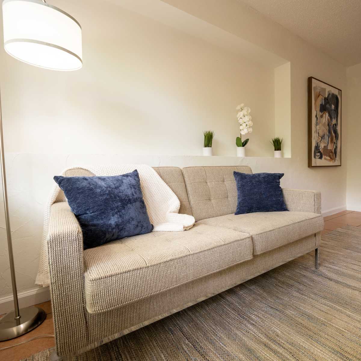 A beige sofa with blue pillows is positioned in a well-lit room, leading to an adjoining area with a visible bed and artwork on the walls.