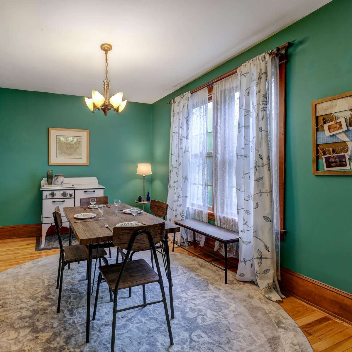 A dining area with a round table surrounded by metal chairs, a wooden cabinet against the wall, and windows adorned with light curtains in a room with green walls.