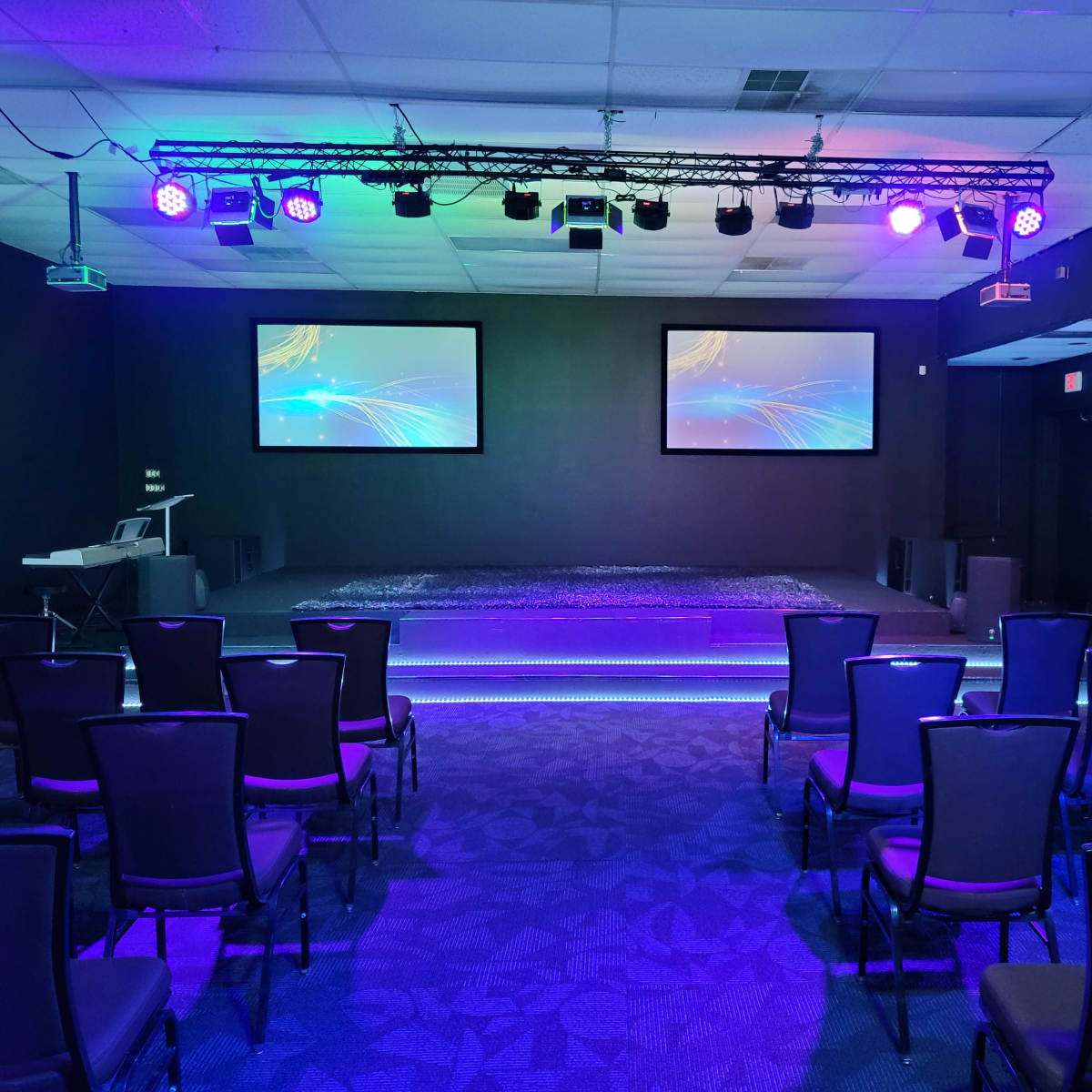 A stage area with two projection screens and purple lighting is set up in a room filled with rows of empty chairs.