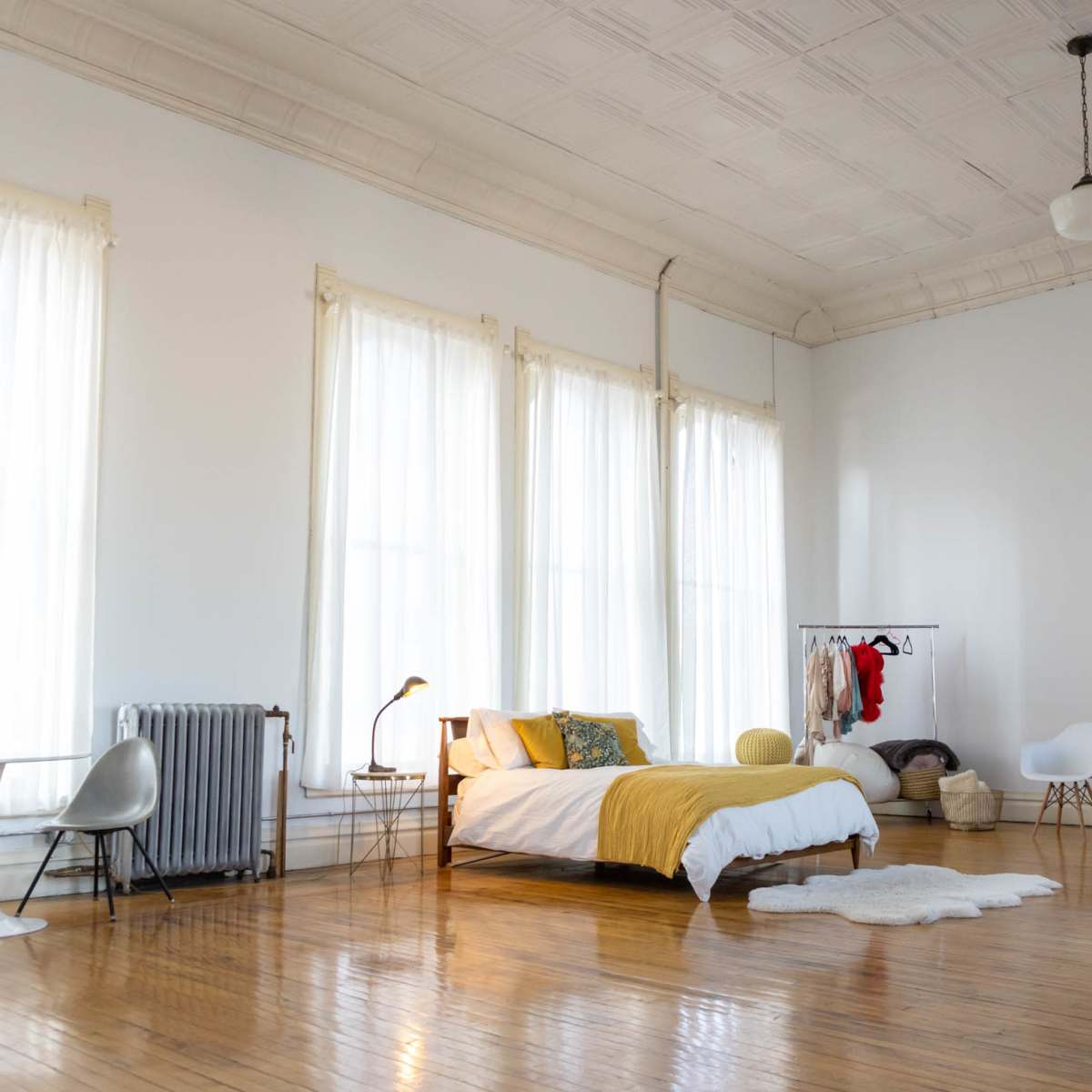 A spacious bedroom features a bed with yellow bedding, large windows draped in sheer curtains, and minimalist furniture including a table and various chairs.