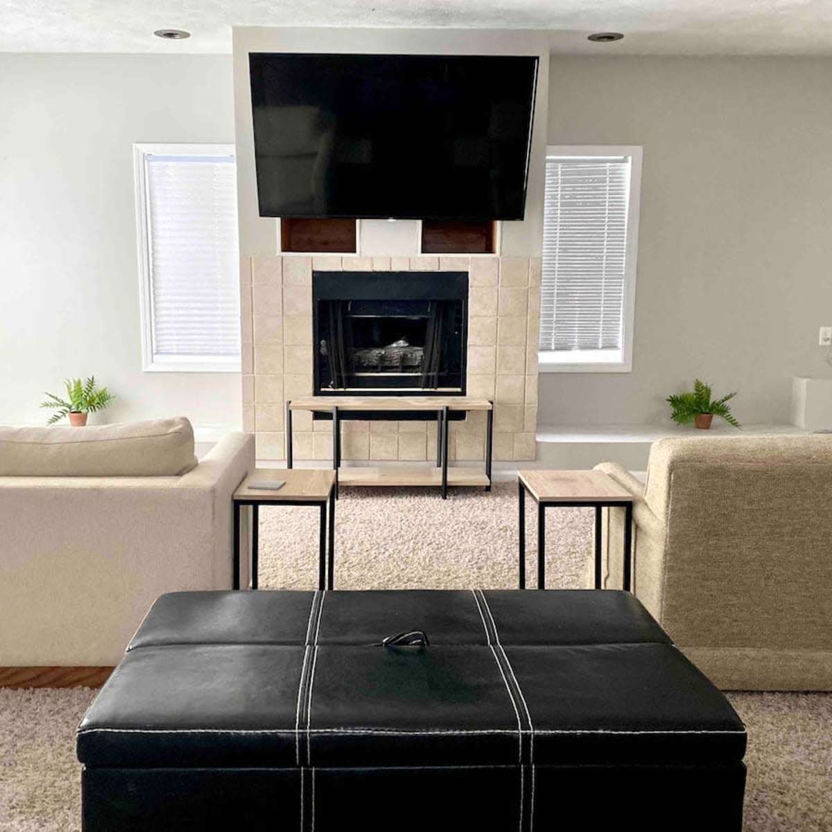 A modern living room with a fireplace, a large television mounted above it, and two sofas facing each other, with a black storage ottoman in the center.