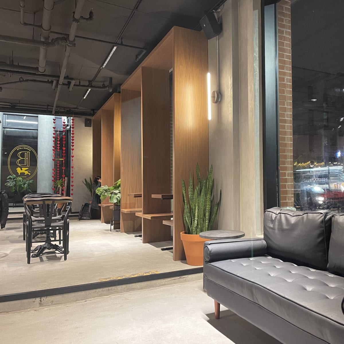 A modern interior of a café with a black sofa, wooden partitions, indoor plants, and large windows reflecting nighttime street activity outside.