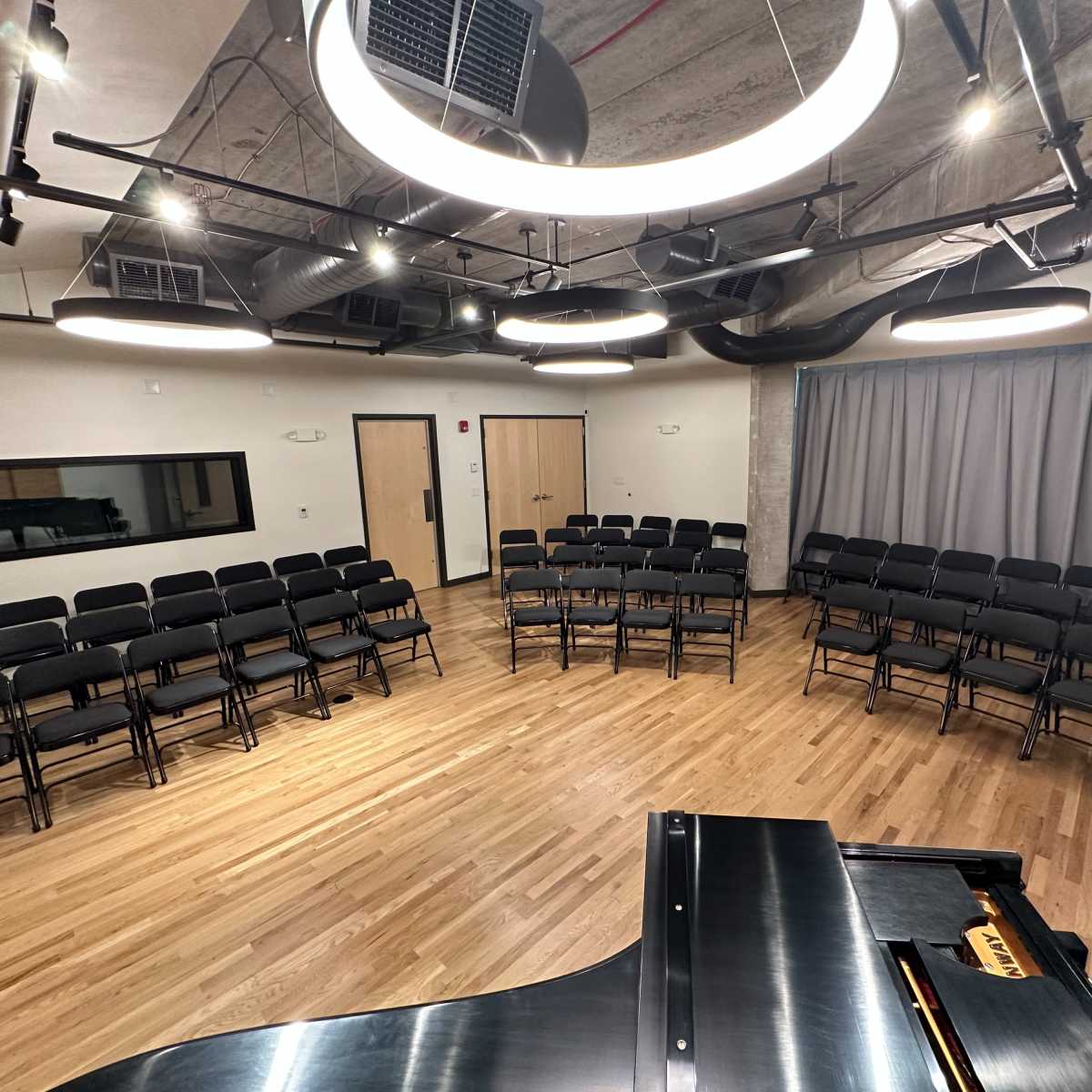 A performance space features rows of black folding chairs arranged in a semicircle around a grand piano, with a curtain and acoustic panels in the background.