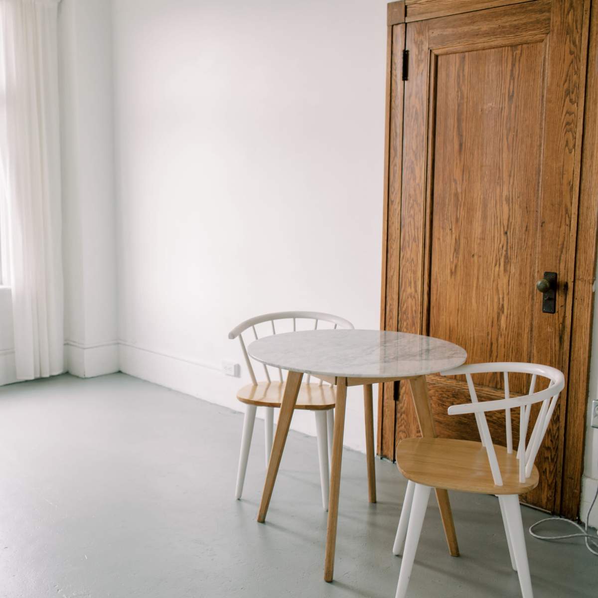 A small, minimalist room features a round marble table with two wooden chairs beside a wooden closet and large windows letting in natural light.