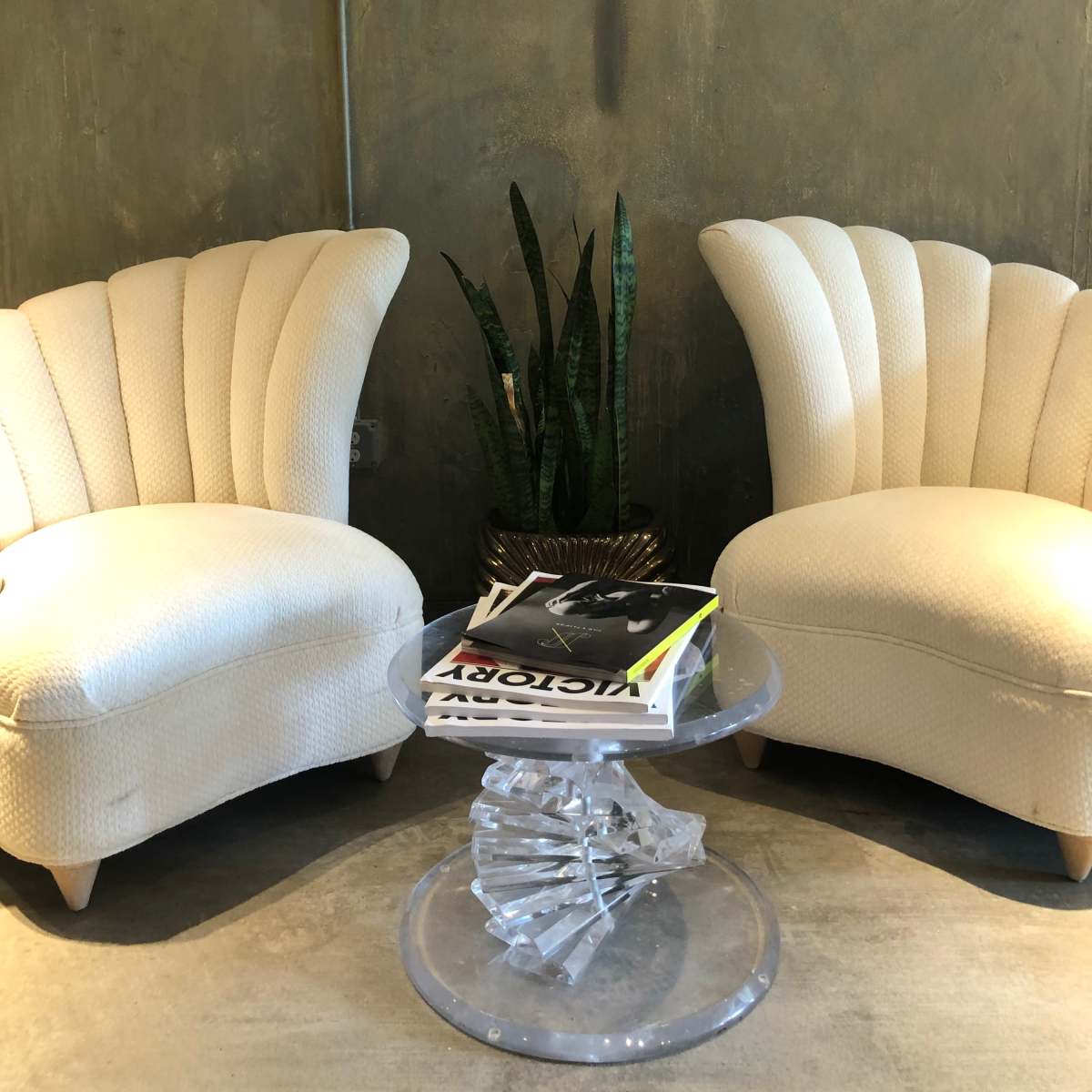 Two white upholstered chairs are positioned facing each other beside a small clear sculptural table, with a potted plant in the background against a textured wall.