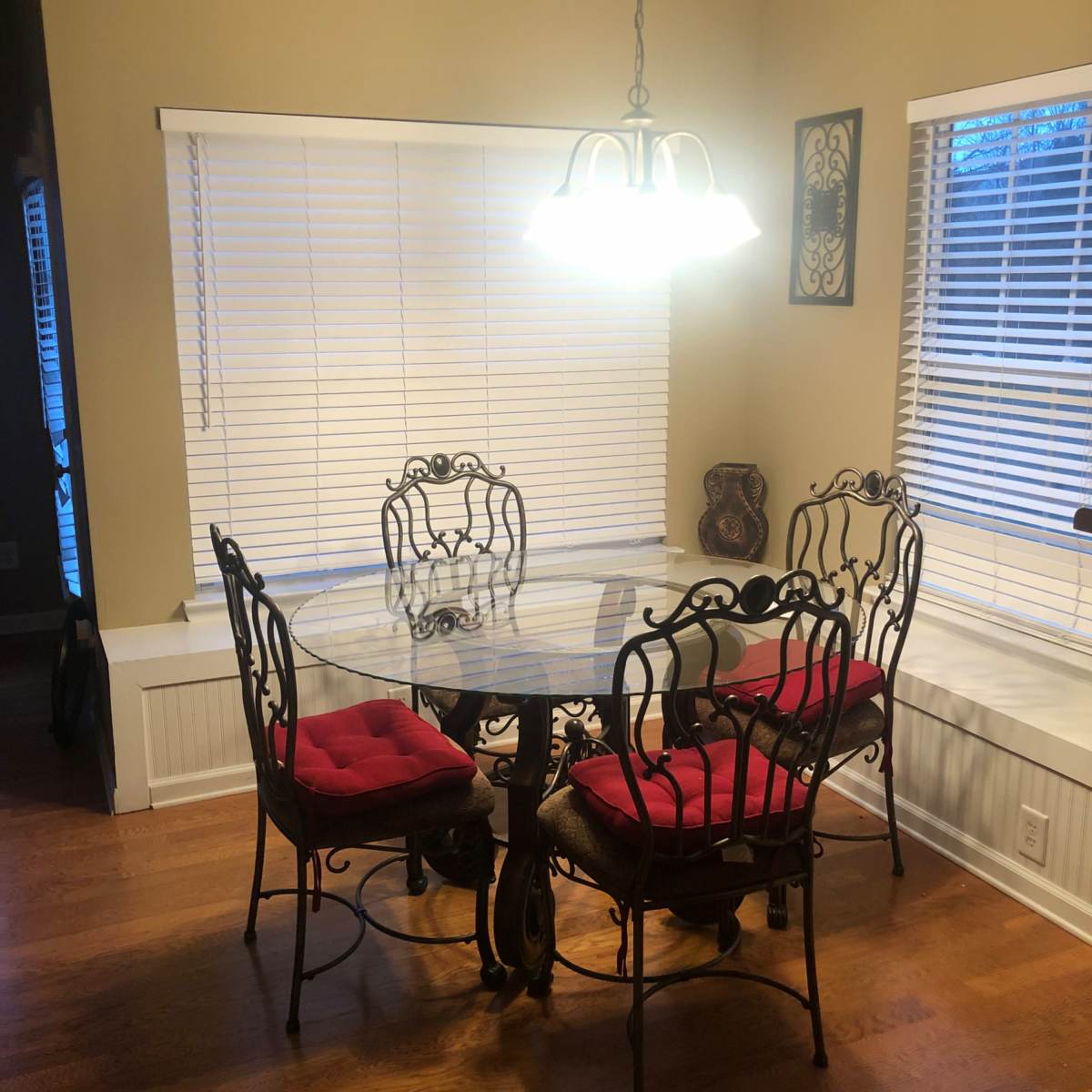 A round glass dining table with four iron chairs, each featuring red cushions, is positioned by a window in a well-lit room.