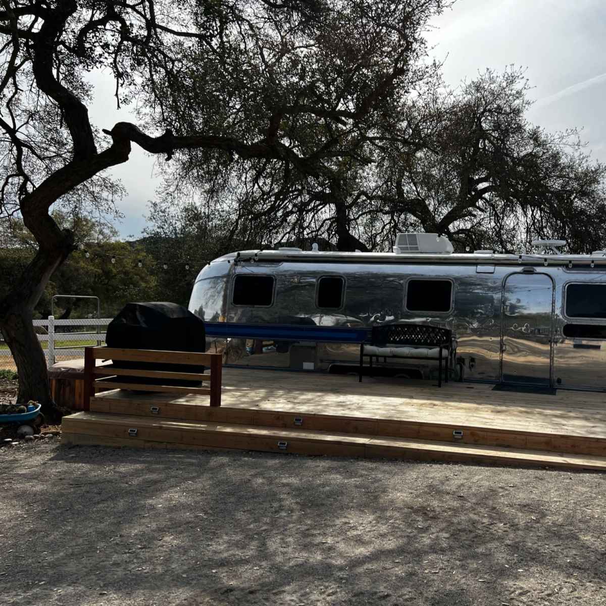 A polished Airstream trailer is parked beside a wooden deck under the shade of an oak tree.