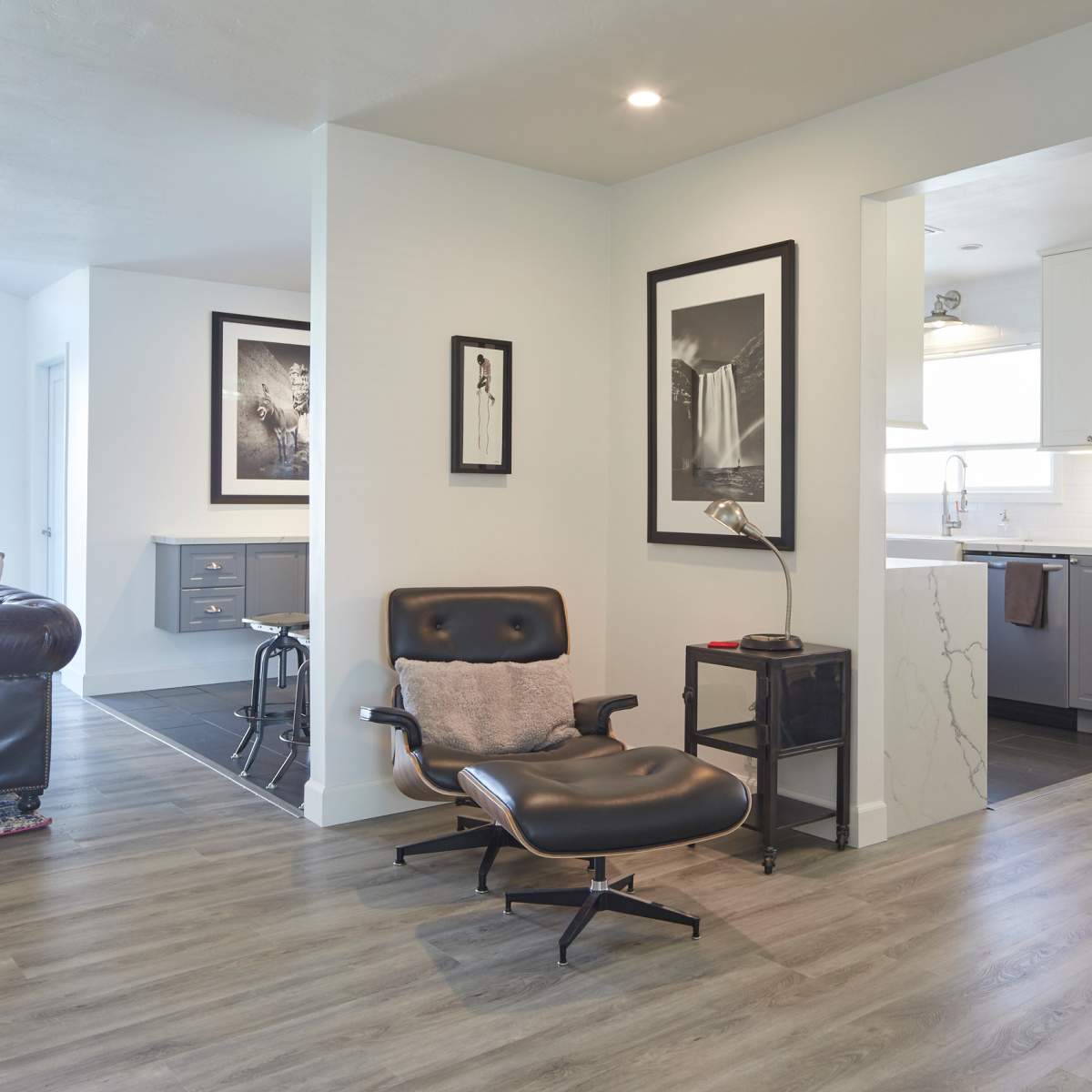 A modern living space features a sleek chair, a small side table, and a transition to a kitchen area with framed artworks on the walls.