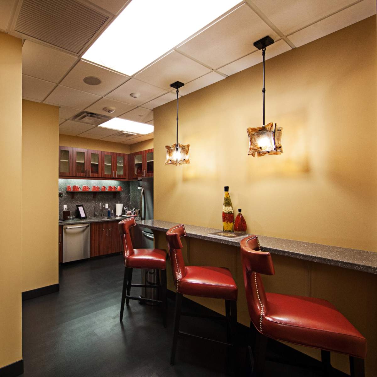 A modern kitchen area with a bar counter and three red stools, featuring cabinets and a small appliance area in a well-lit space.