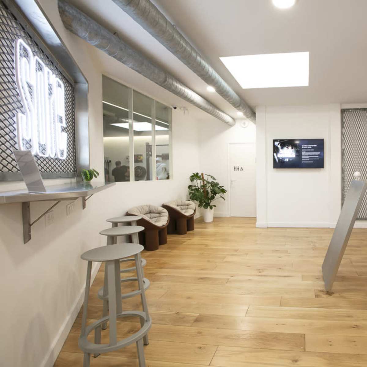 The interior of a modern room features a light wooden floor, a bar-style table with stools, and a television mounted on the wall.