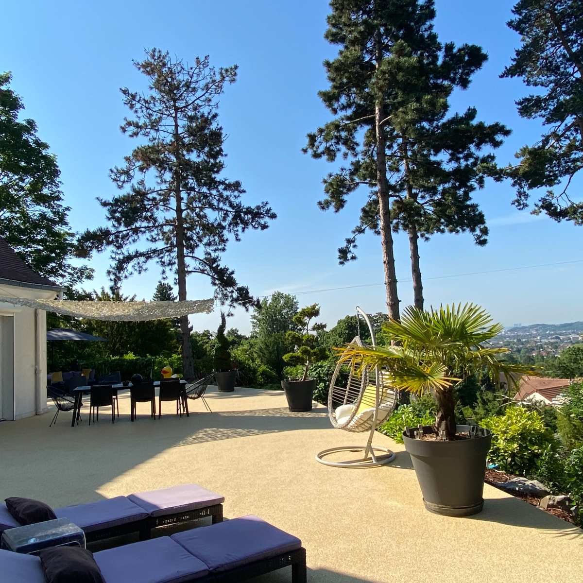 A spacious patio with lounge chairs, a hanging chair, and potted plants overlooks a scenic view surrounded by tall trees.