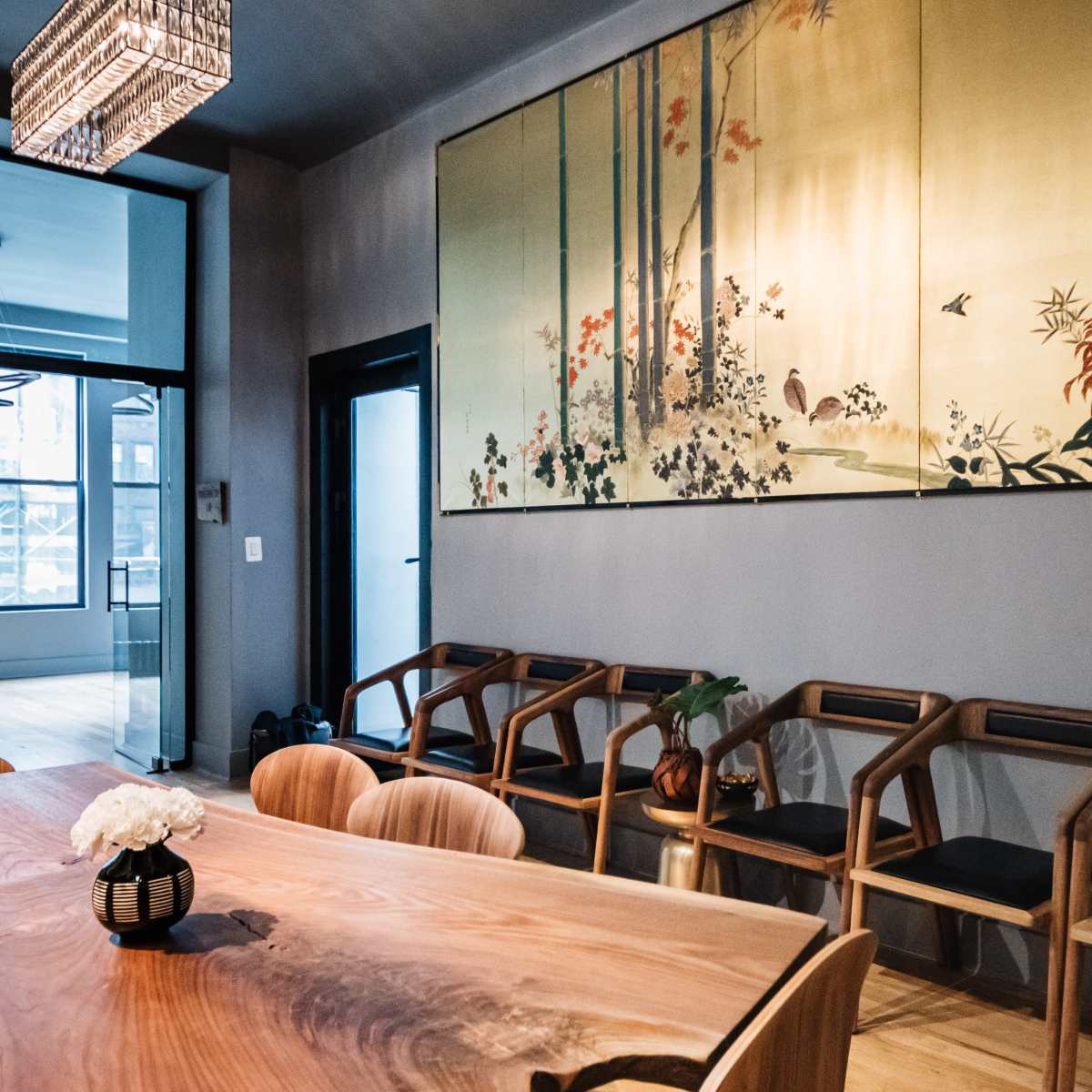 A wooden dining table is surrounded by chairs in a room featuring a large decorative wall hanging of bamboo and flowers.