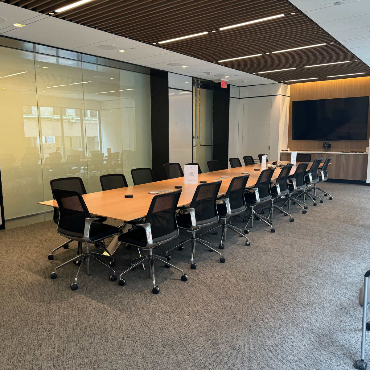 A spacious conference room features a long wooden table surrounded by black office chairs and a wall-mounted screen in the background.