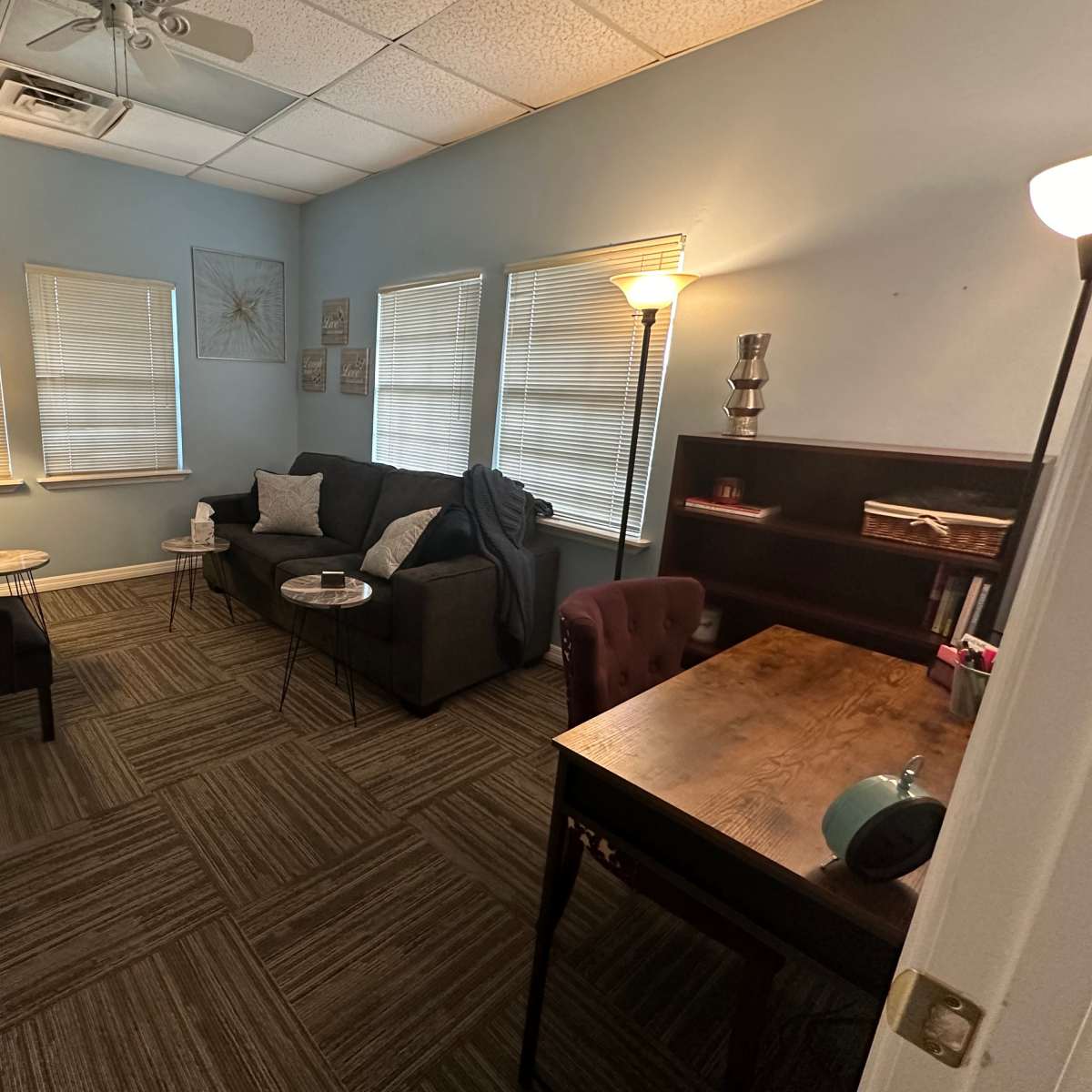 A small office space with a couch, two chairs, a coffee table, a desk, and multiple windows.