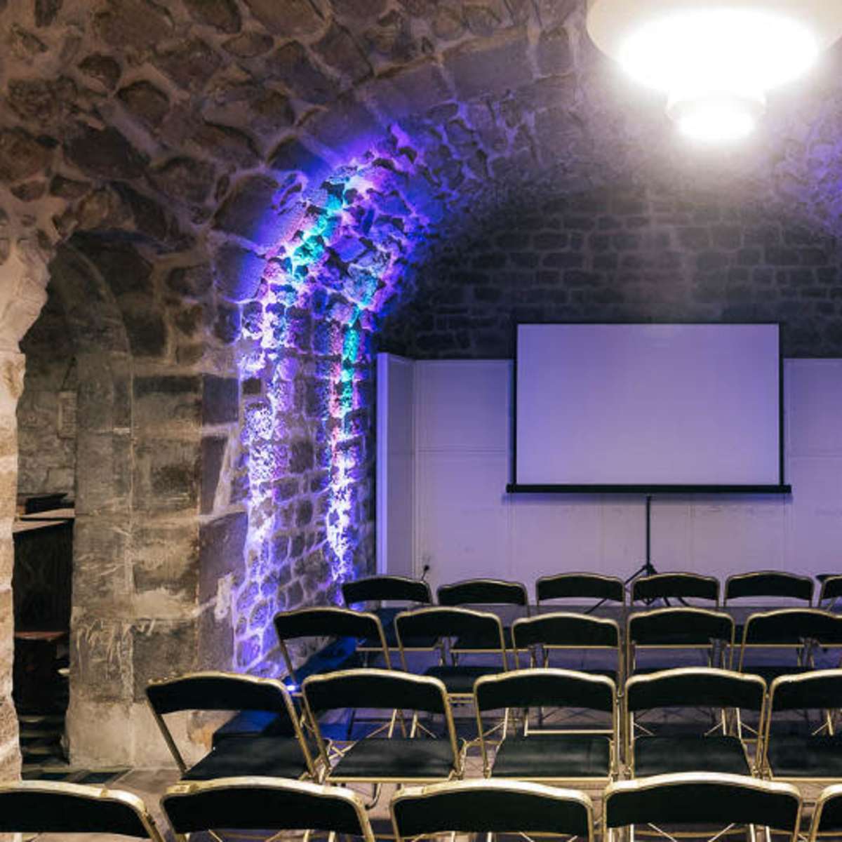 A stone-walled meeting room features rows of black chairs facing a large screen, illuminated by colored lights.