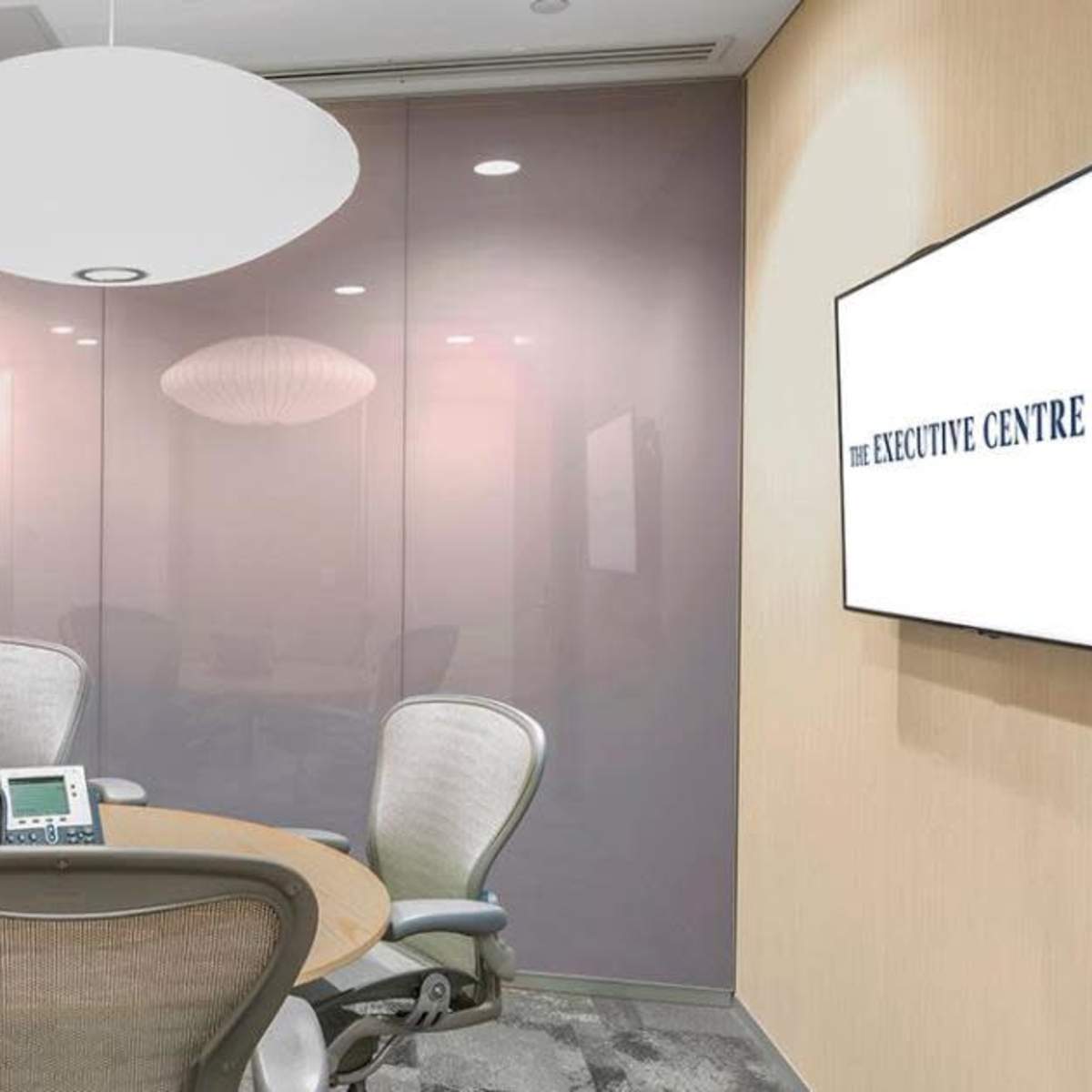 A modern conference room features a round wooden table and ergonomic chairs, with glass walls and a wall-mounted screen displaying "THE EXECUTIVE CENTRE."
