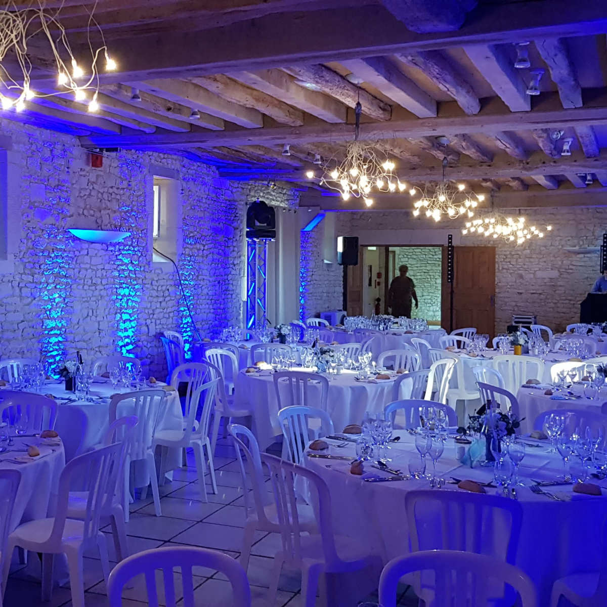 A banquet hall set with round tables covered in white tablecloths, illuminated by blue lighting from the walls and ceiling fixtures.