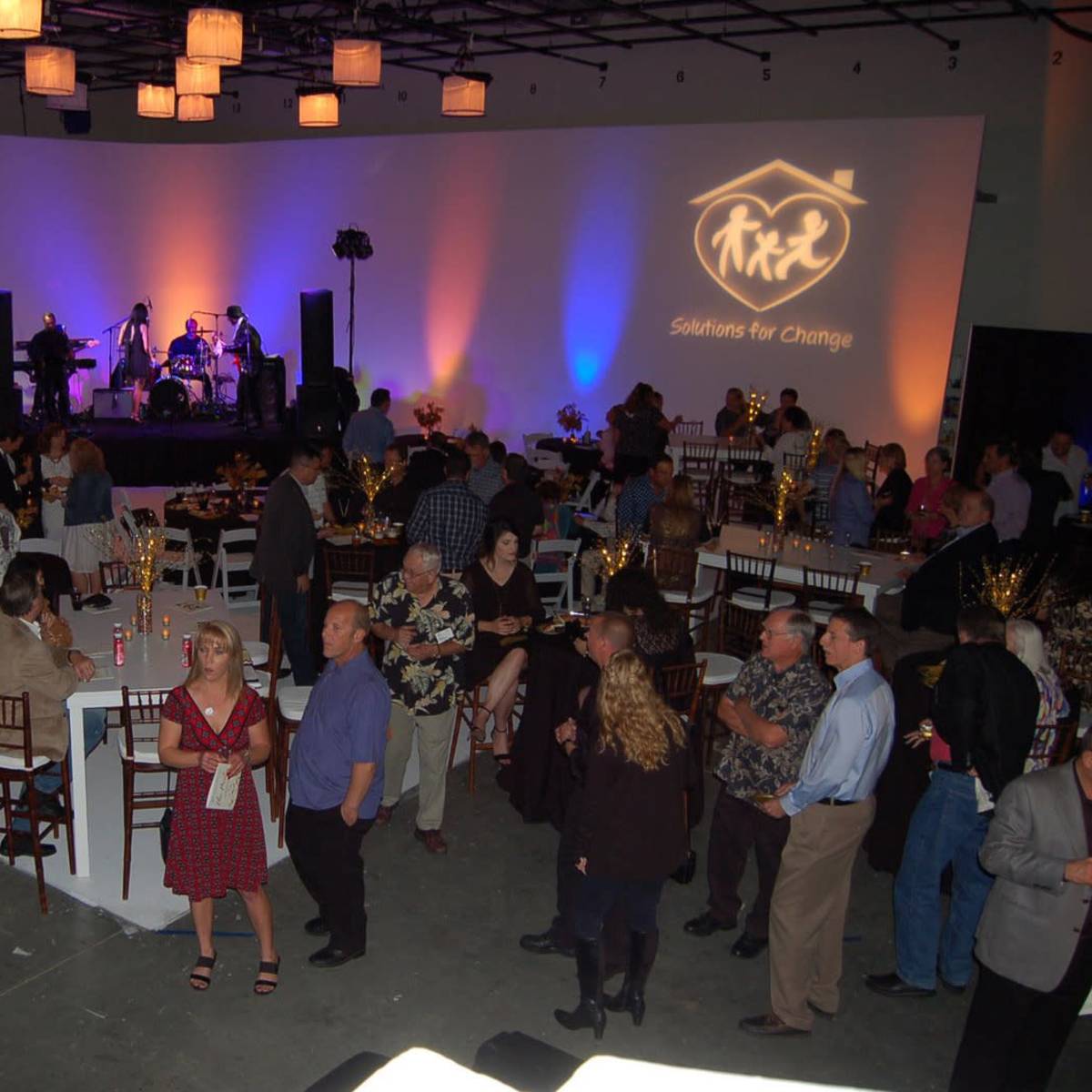 A crowd gathers at a gala event featuring a live band on stage, with tables set for dining under decorative lighting.