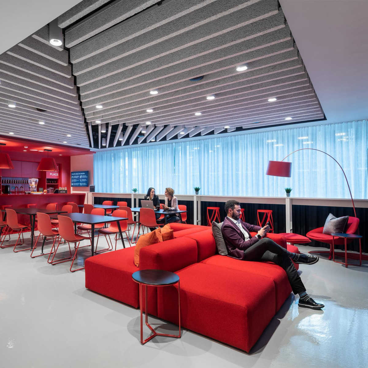 A modern lounge area features red seating, a bar counter, and three people engaged in conversation or relaxation.