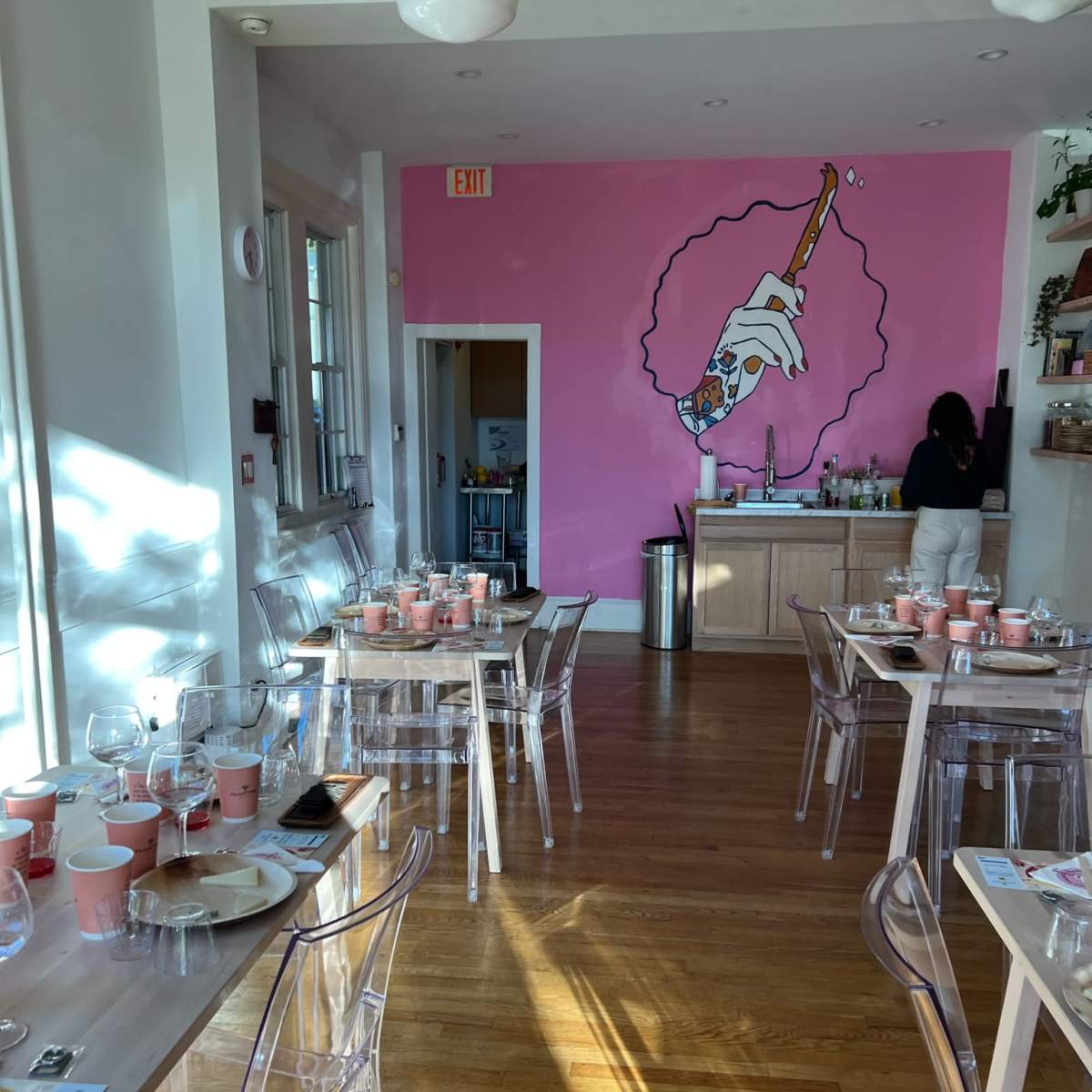 A brightly lit restaurant features transparent chairs, tables set with cups, and a large pink mural of a hand holding a cigarette on the wall.