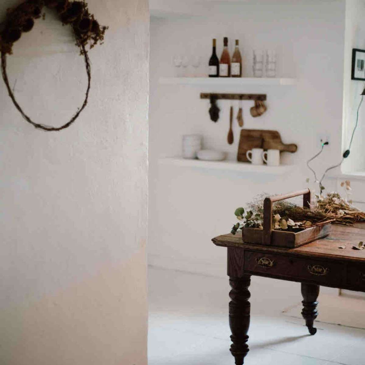 A wooden table with a rustic centerpiece is positioned in a softly lit kitchen that features white walls and open shelving with dishes and bottles.