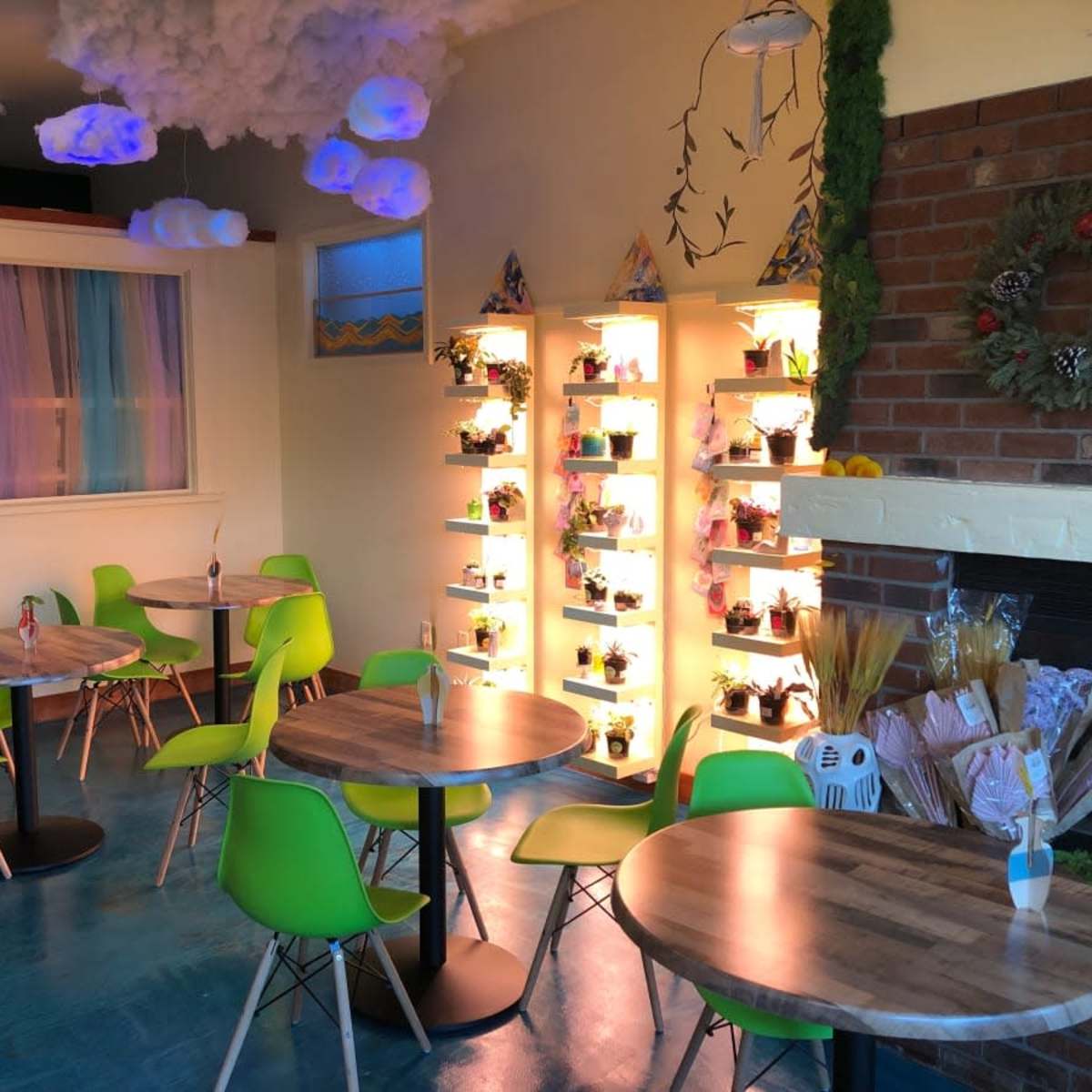 A colorful cafe interior with green chairs and small wooden tables, lit by decorative shelves filled with plants and illuminated by soft hanging clouds.