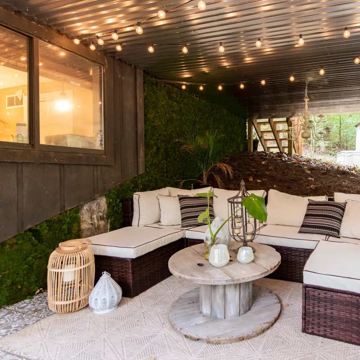 A covered outdoor seating area features a sectional sofa with beige cushions, a round wooden coffee table, and decorative string lights overhead.