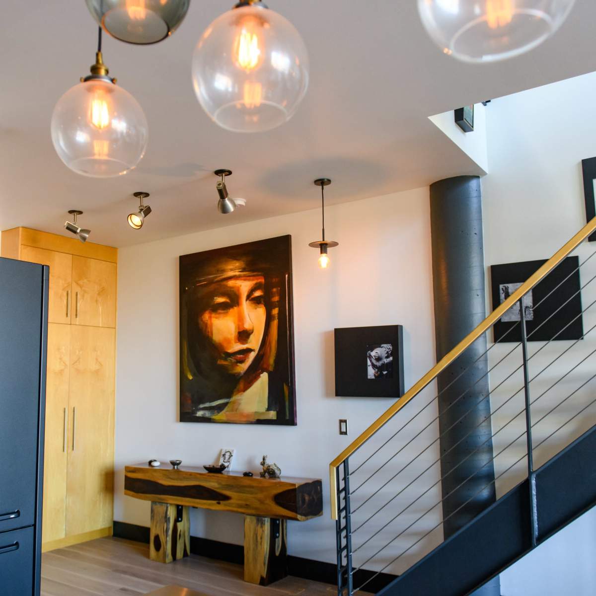 A modern interior space with a staircase, a large painting of a woman on the wall, wooden furniture, and various light fixtures hanging from the ceiling.