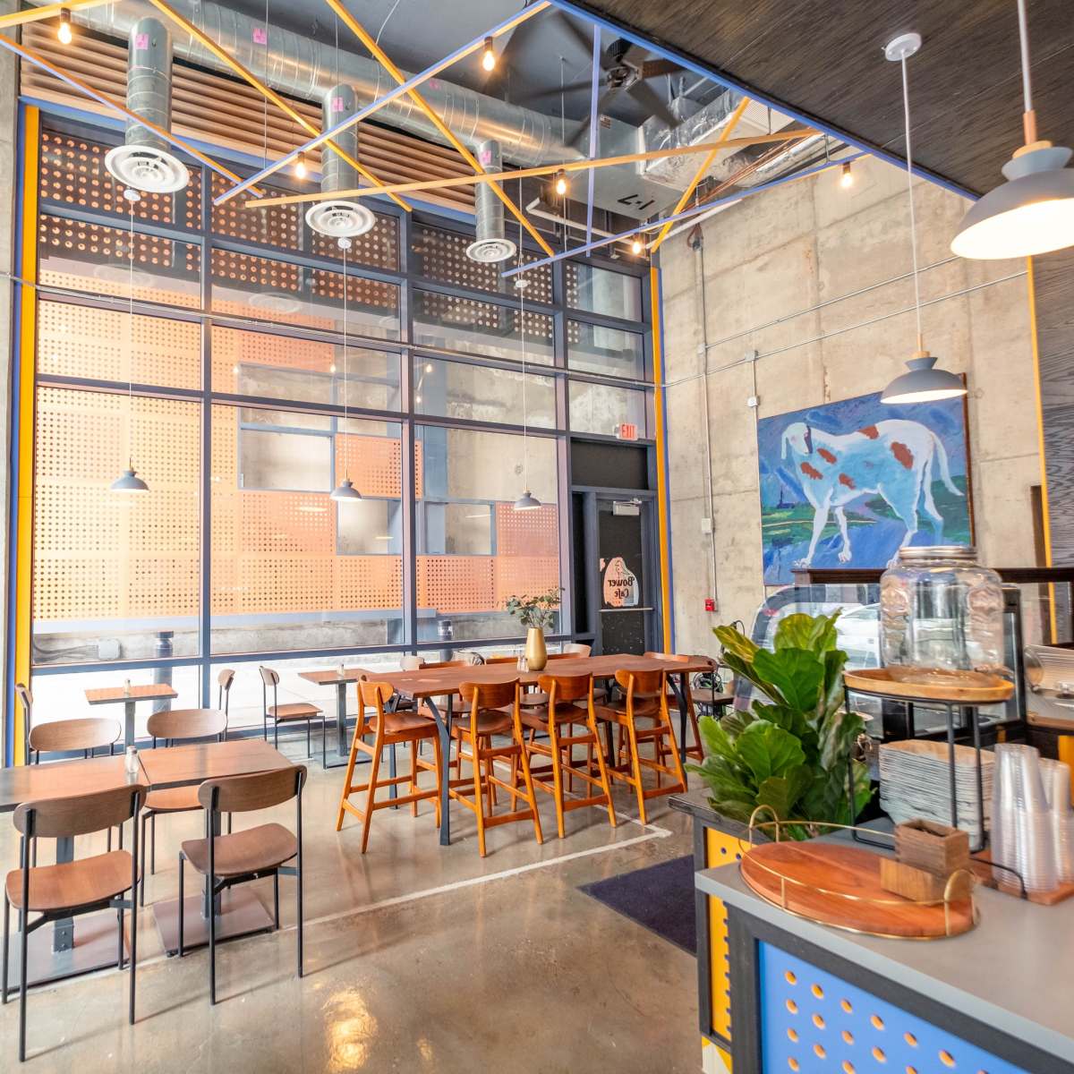 A modern cafe interior with metal and wooden furniture, large windows, and a colorful wall art piece.