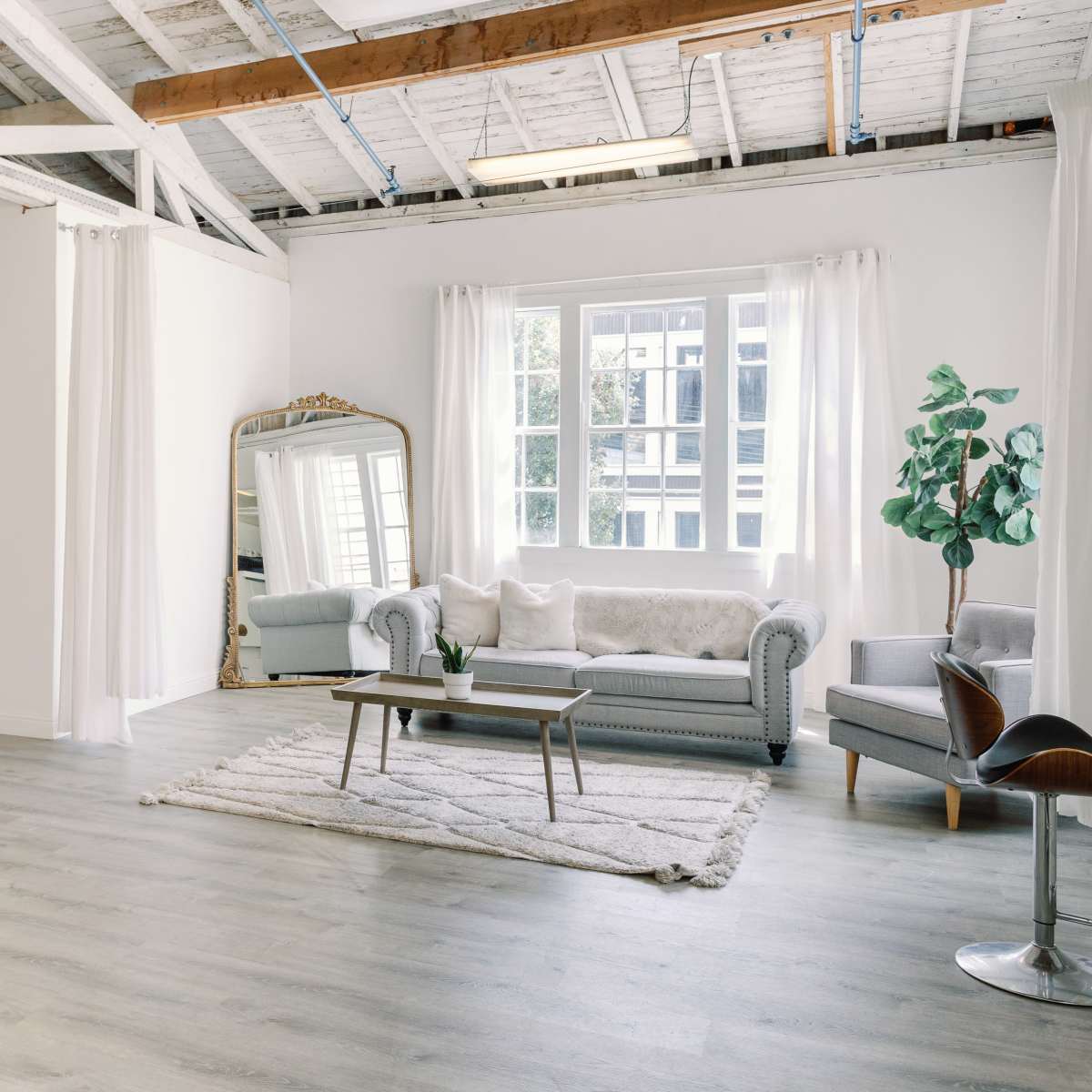 A spacious, bright living area with light-colored wooden flooring, a large window, a gray sofa, a mirror, and various decorative furniture items.