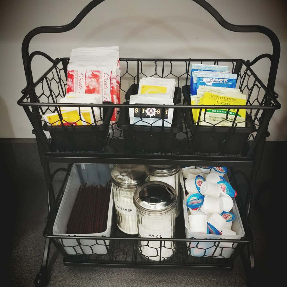 A black wire rack contains various packets of tea, sugar, and creamers on its upper shelf, while the lower shelf holds glass containers for sugar and a box of stirrers.