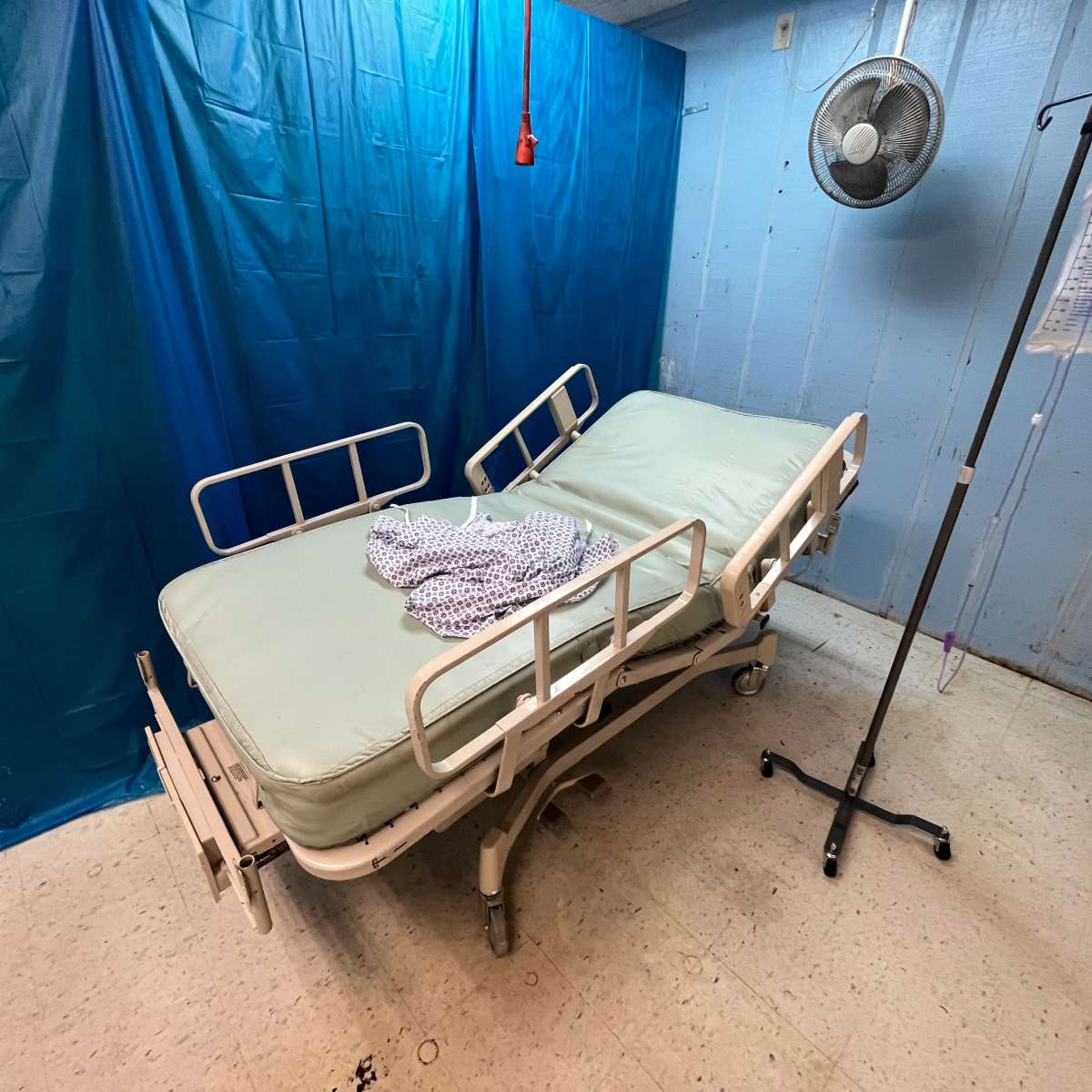 A hospital room with an adjustable bed, a fan, and an IV stand beside it, all set against a blue backdrop.