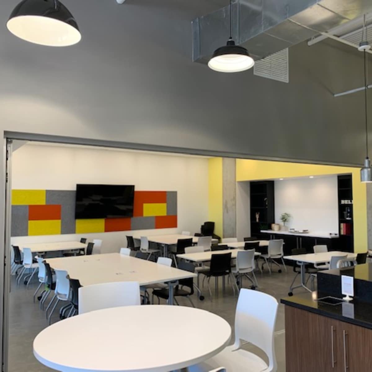 A modern, open-plan dining area with white tables and chairs, a large television screen on the wall, and a neighboring workspace featuring shelves and plants.