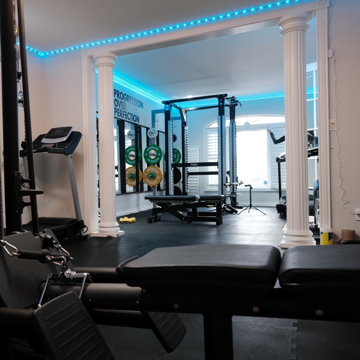 A well-equipped home gym featuring weightlifting machines, exercise mats, and training equipment, with blue LED lighting and columns in the interior design.