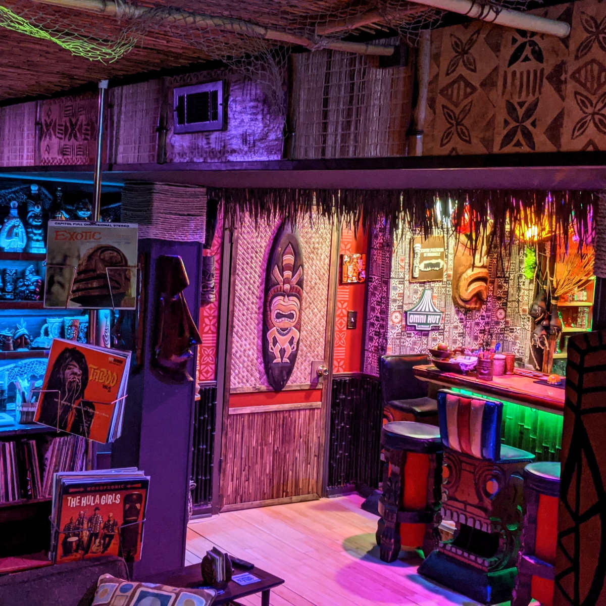 A dimly lit tiki bar with colorful lighting, featuring a wooden counter, tiki masks on the walls, and shelves filled with tropical-themed decor and vintage records.