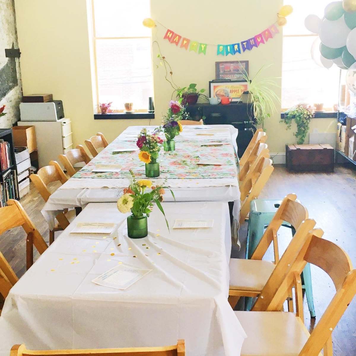 A decorated indoor space with two long tables covered in white tablecloths and surrounded by wooden chairs, featuring colorful birthday decorations and flower centerpieces.