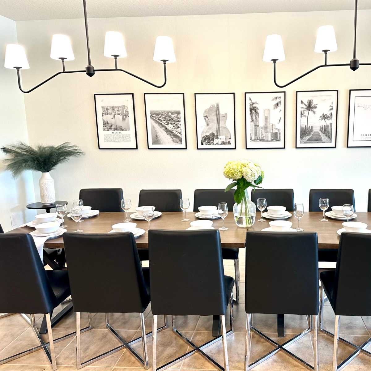 A long dining table is set with plates, glasses, and a vase of flowers, with eight black chairs arranged on each side and six framed pictures hung on the wall above it.