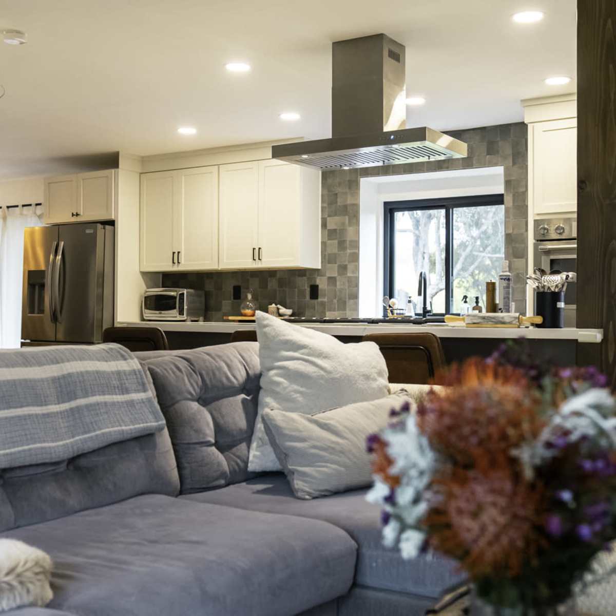 A modern kitchen is visible in the background, featuring stainless steel appliances and white cabinetry, with a cozy living area in the foreground that includes a gray couch and decorative flowers.