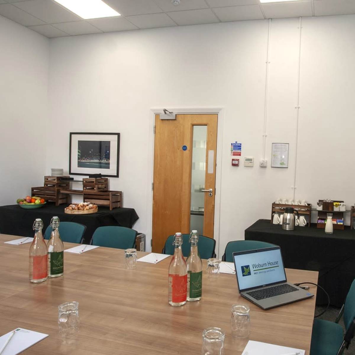 A conference room is set up with a wooden table, chairs, and presentation equipment, featuring a side table with refreshments and an entrance door.