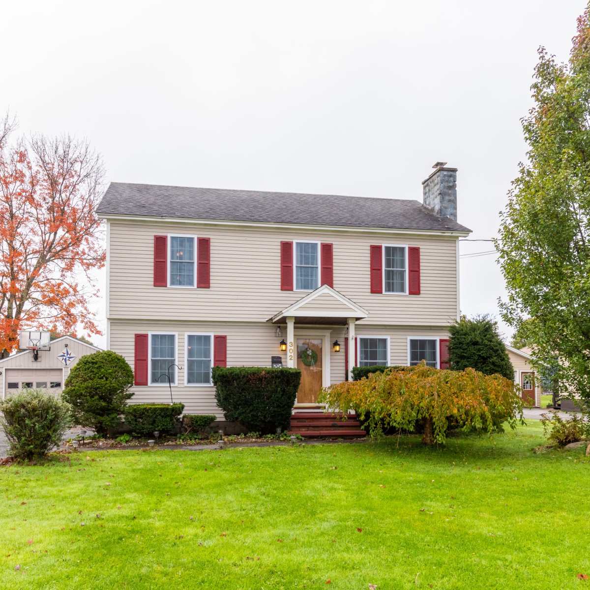 A two-story house with red shutters sits on a green lawn, surrounded by trees and a gravel driveway.