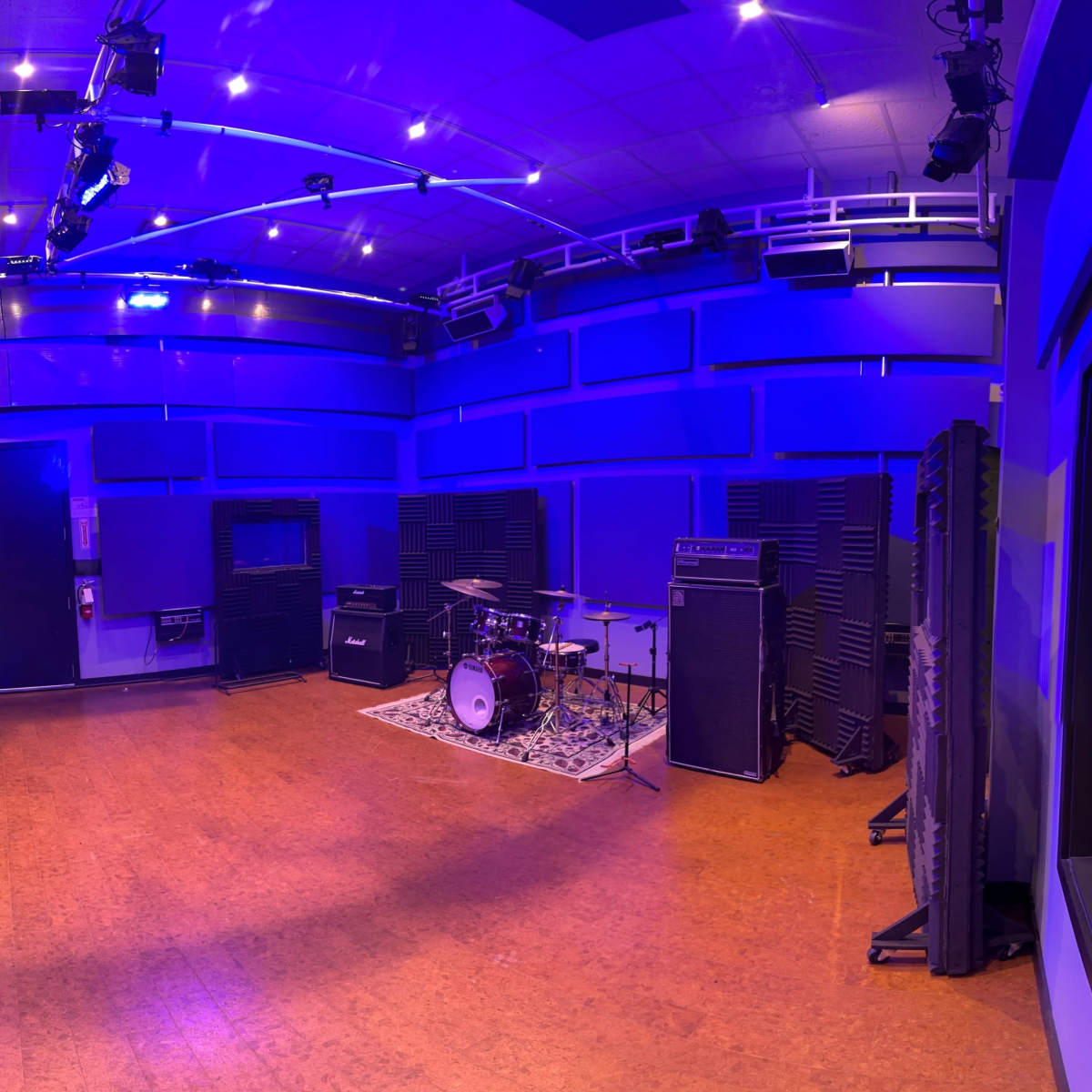 A sound studio with a drum kit and amplifiers in a blue-lit room, viewed through a glass panel from an adjacent control room.