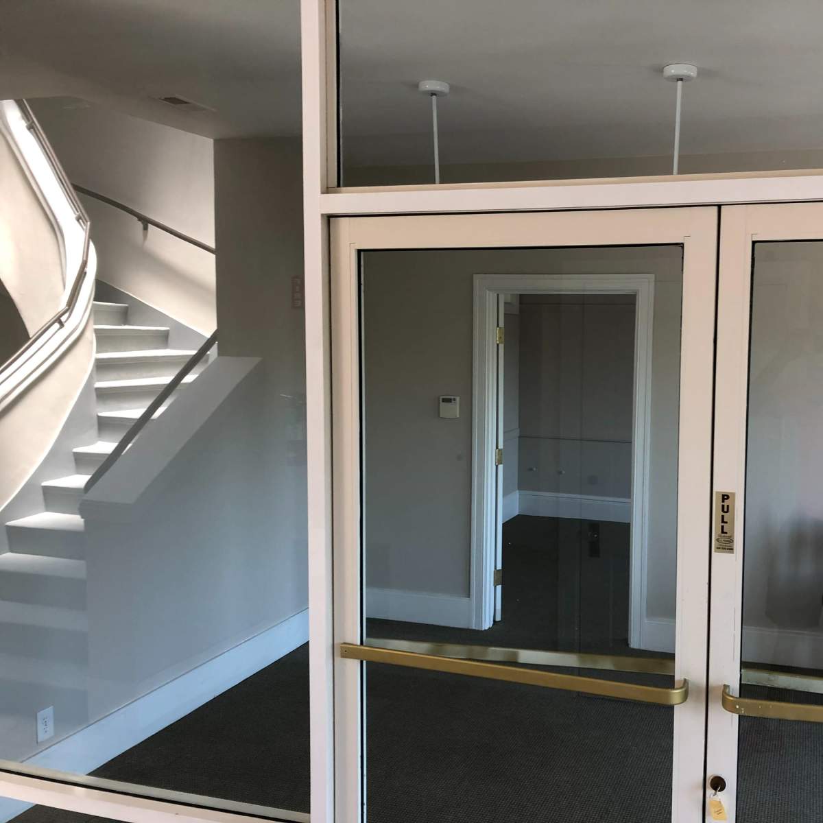 A glass entrance with a reflection of a staircase and a room featuring several stacked chairs behind it.