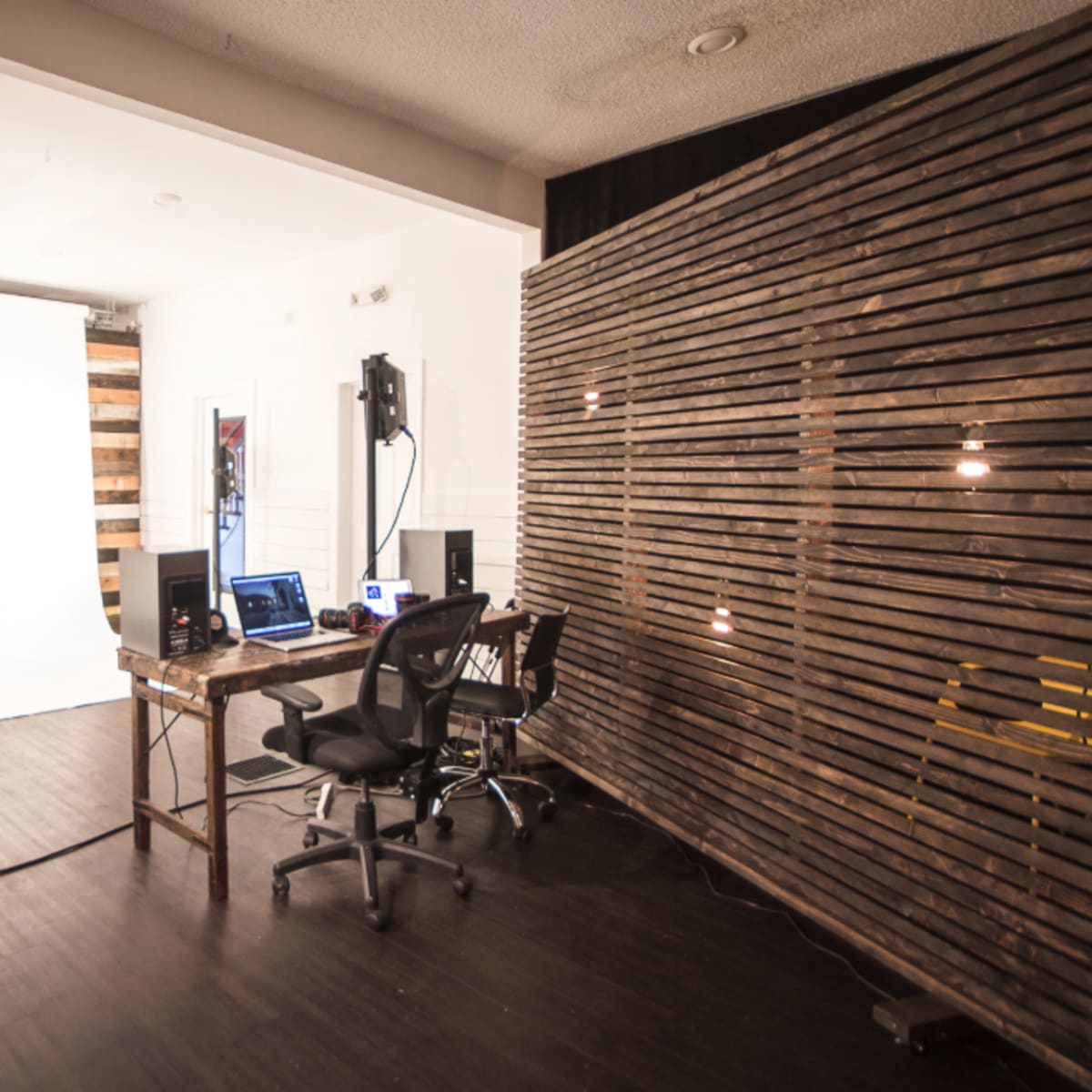 A photography studio with a white backdrop, lighting equipment, and a workspace featuring a wooden table and chairs.