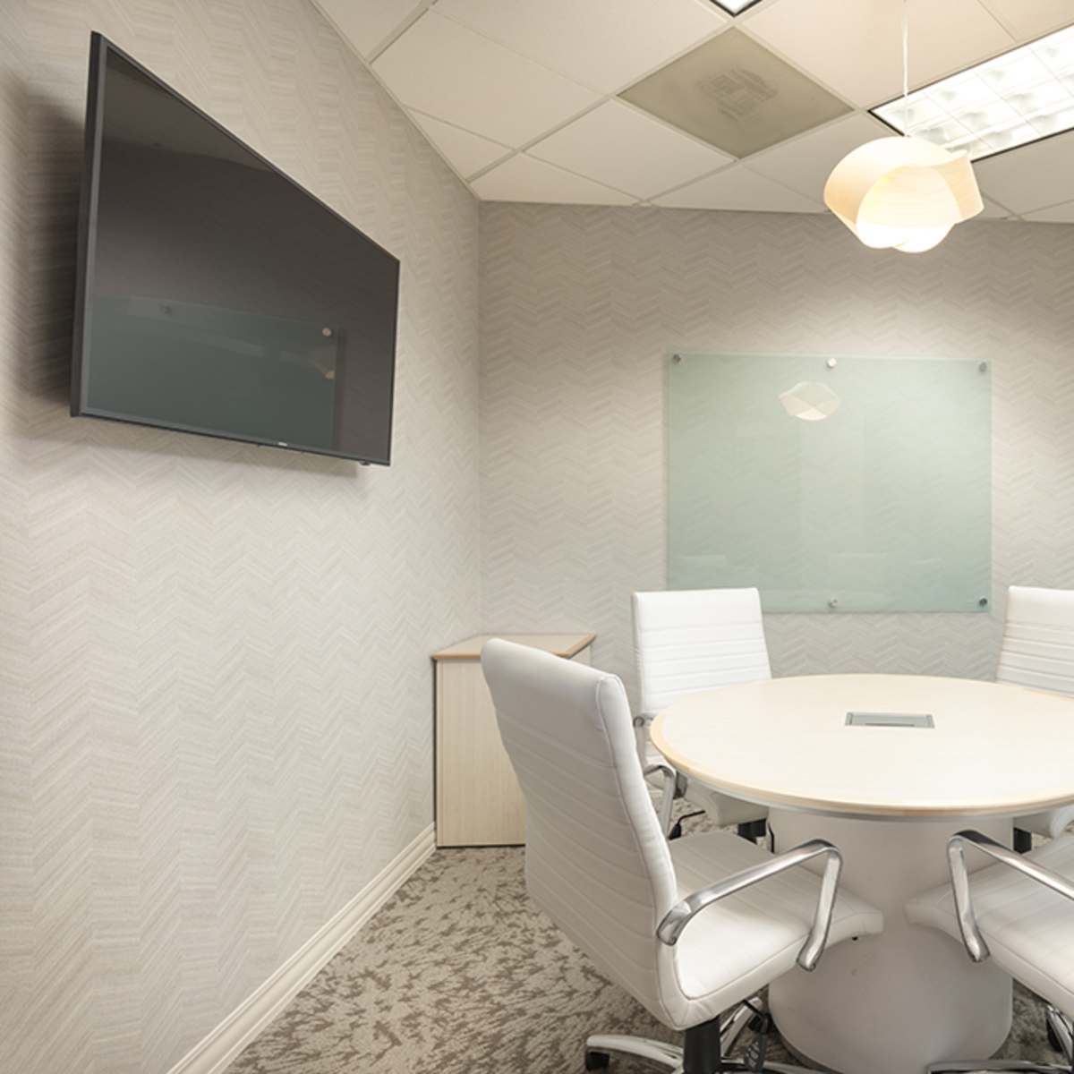 A modern conference room with a round table, four white chairs, a wall-mounted TV, and a glass partition.