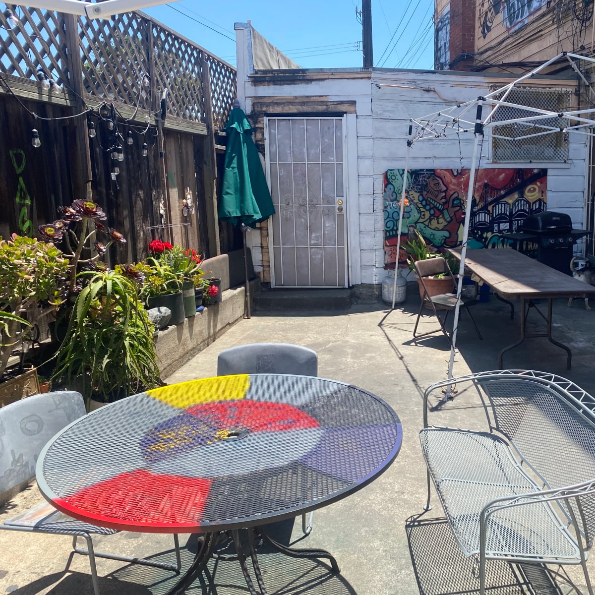 A small outdoor patio area with a round table, several chairs, a weathered shed, and a canopy frame set up beside colorful murals and potted plants.