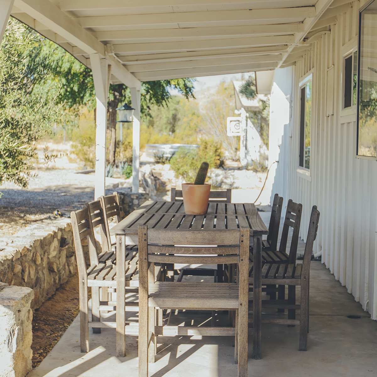 A wooden dining table with matching chairs sits under a covered porch, accompanied by a potted cactus.