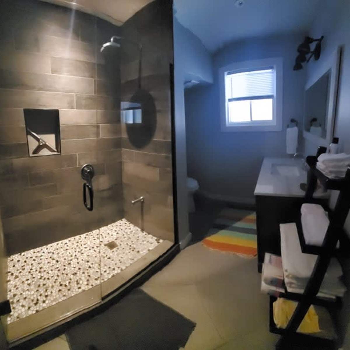 A modern bathroom with a glass shower enclosure featuring stone tile and a pebble-style floor, alongside a vanity area and a small window.