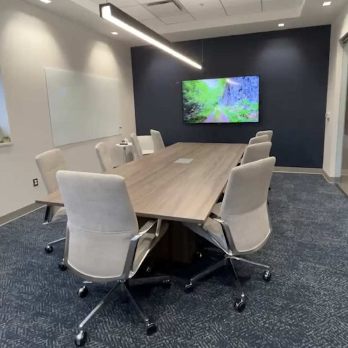 A modern conference room featuring a long table surrounded by ergonomic chairs and a large screen displaying a nature scene on the wall.