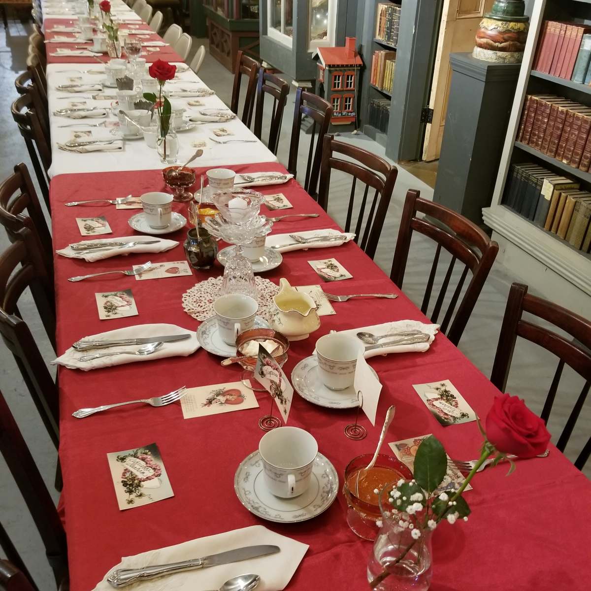 A long dining table is elegantly set with white china, silverware, and decorative flowers, all arranged on a red tablecloth.