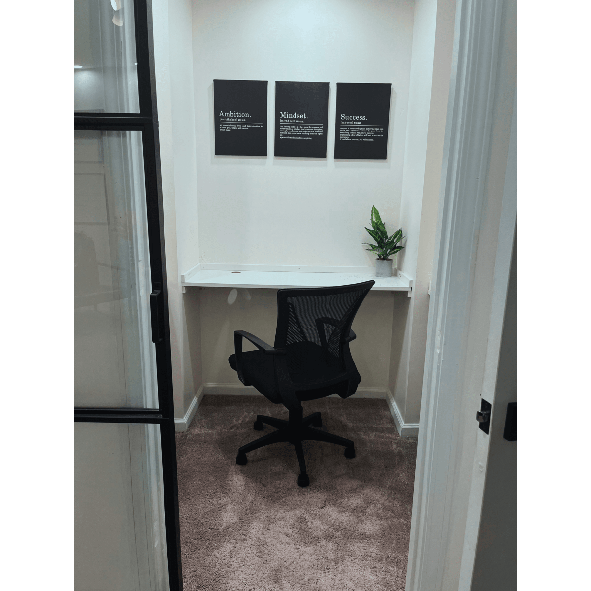 A small office space features a desk with an ergonomic black chair, three framed motivational quotes on the wall, and a potted plant.